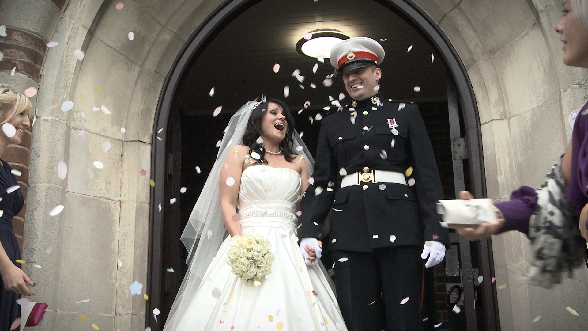 a groom in military dress and bride in white laugh as they stand in their wirral church doorway being showered in confetti for the wedding video