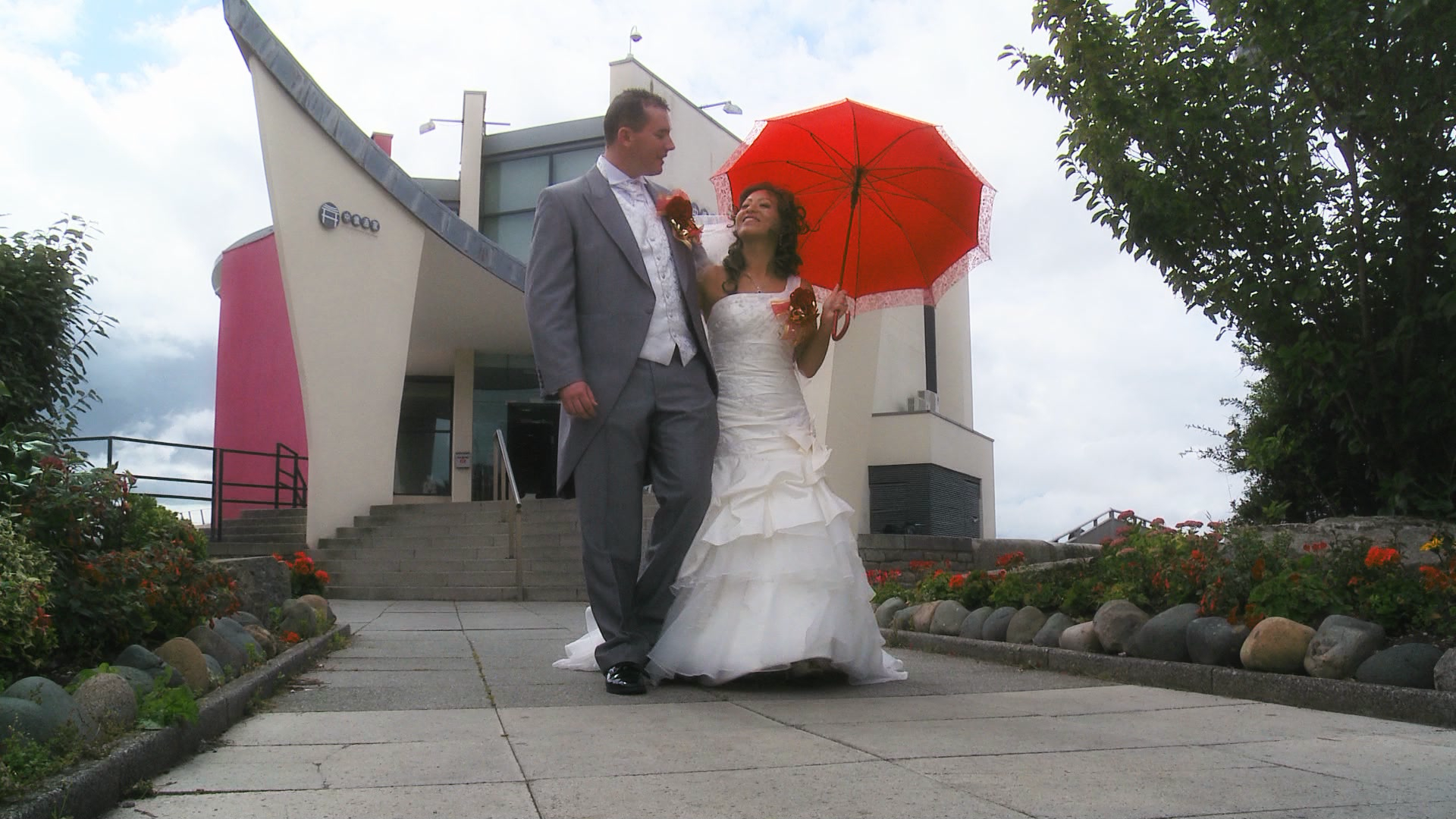 A bride and groom walk towards the videographer as the bride holds a traditional red chinese umbrella before their wedding reception at Chung Ku by the liverpool waterfront
