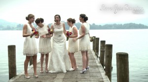 A bride and her bridesmaids hold each other shocked on a jetty in Lake Windermere after one of the bridesmaids nearly fell in due to her heel getting stuck and theyre laughing towards their wedding videographer