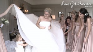 A beautiful bride stands in the bridal suite at Thornton Manor with her bridesmaids wearing pink two bird bridesmaid dresses. They're helping her get ready as a cute toddler flower girl goes under her dress