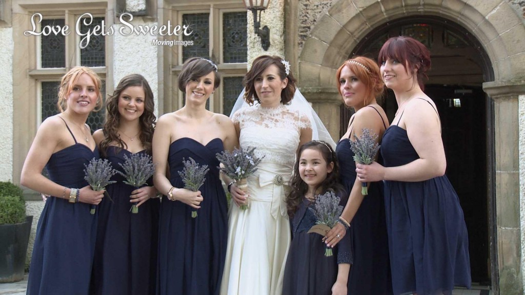 a bride wearing stewart parvin lace and silk wedding dress holding lavender faux flowers poses with her bridesmaids wearing floor length navy blue coast dresses as Mick Cookson takes photos and love gets sweeter video outside Mitton Hall