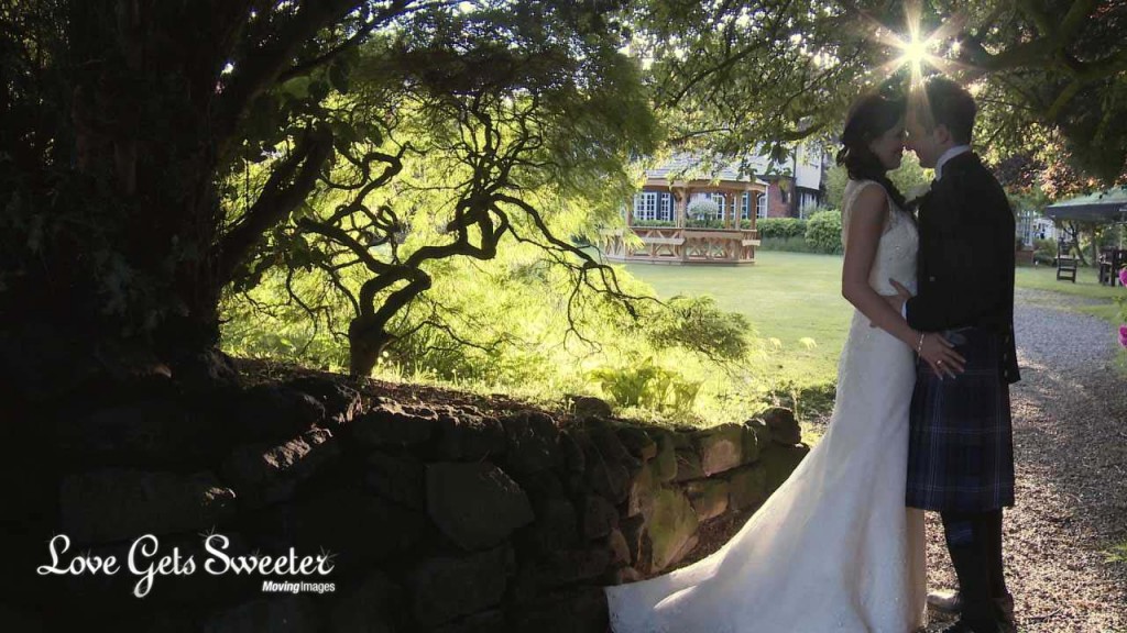 the bride and groom enjoy a moment together with the wedding videographer under the trees at Mere Court Hotel with the sun creeping through the branches. She wears a lace wedding dress and he wears a traditional Scottish kilt