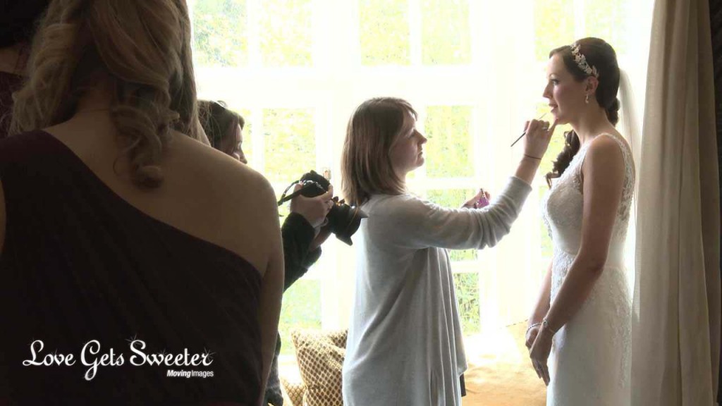 the bride stands by a bright window in a suite at the Mere Court Hotel near Knutsford as her wedding makeup artist applies her lipstick. This is a video still from Love Gets Sweeter