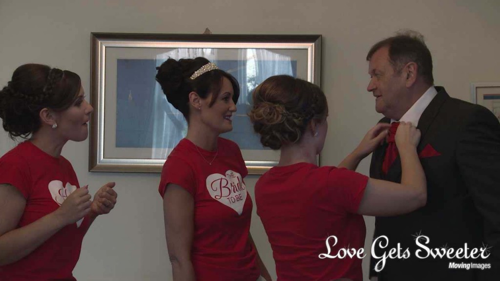 A bride and her sisters wear matching red wedding t-shirts as one of them helps straighten their dads red wedding tie in their room in West Tower