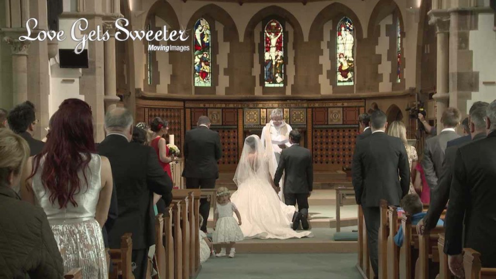 a video still from the back of Dalton Church as the bride and groom kneel during their wedding ceremony for the blessing. A cute flower girl wanders around the aisle
