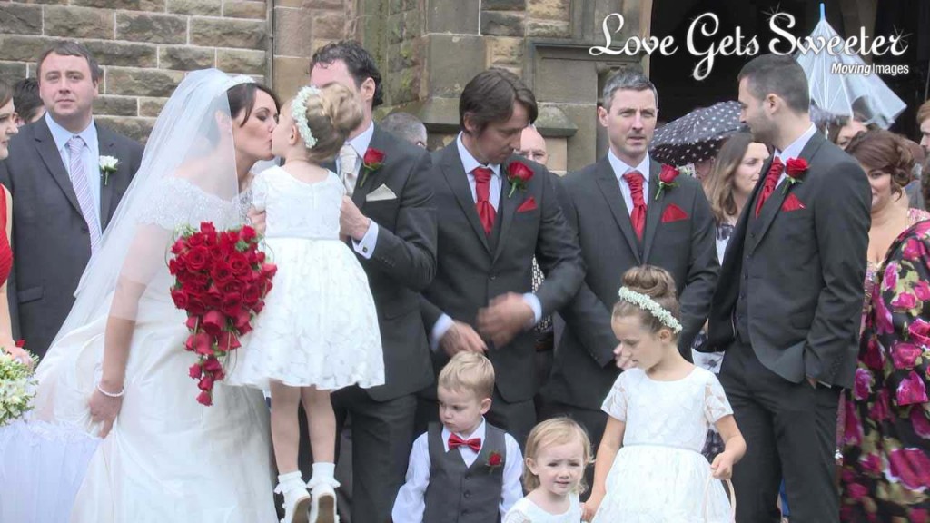 The groom lifts up one of their daughters during the wedding photographs so the bride can have a kiss. She holds a traditional heart shaped red rose bridal bouquet and the wedding party wear red ties for their white and red themed wedding
