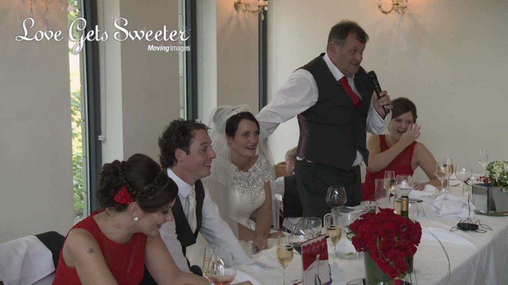 A wedding video still during the wedding speeches of a bride looking mortified during the father of the bride speech at their West Tower wedding in Aughton Lancashire. Bright red roses add to the red and white colour scheme.