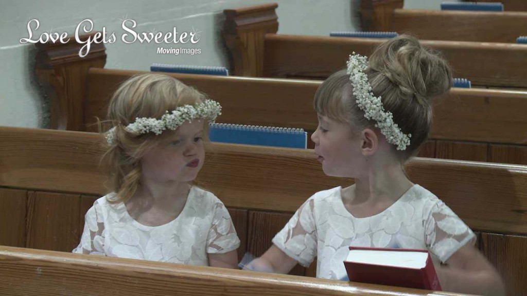 2 very cute flower girls wearing white gypsy grass flower crowns sit in a pew at Dalton Church. One is pouting at the other