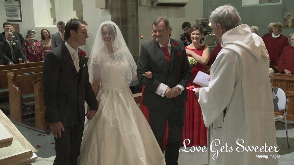 a very proud dad looks on as his daughter meets her groom at Dalton Church with her veil still over her face. Her bridesmaids stand to the side wearing floor length red bridesmaid dresses