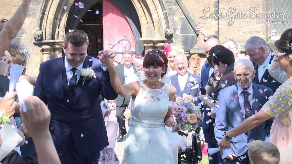 a wedding video still of the bride and groom holding hands as they get showered in confetti by their wedding guests outside their church in New Brighton