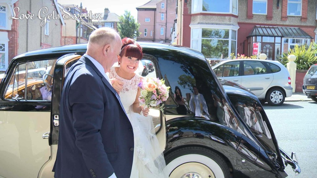 A still from Siobhan and Gary's wedding video of the bride stepping out on her cream and black traditional wedding car in to the sunshine with a huge smile on her face holding a bright pastel bridal bouquet in the Wirral