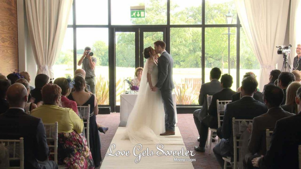 a still from their wedding video of a bride and groom kiss as their wedding guests clap and cheer after being announced husband and wife during their wedding ceremony at Rookery Hall in Nantwich