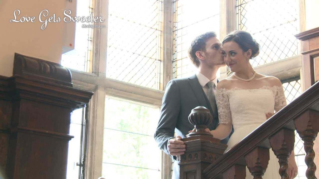A groom kisses the cheek of his bride as they stand on the dark wooden staircase at Rookery hall to pose for the photo and video. She wears an off the shoulder lace vintage inspired wedding dress and pearl neckalace