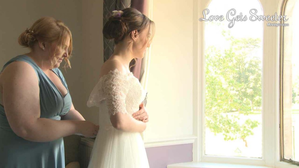 the bride stands in front of a huge window in a suite at Rookery Hall as a bridesmaid wearing blue helps her in to her off the shoulder lace vintage inspired wedding dress for her wedding video