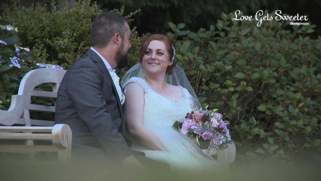 A still from wedding videographer Love Gets Sweeter of a bride and groom enjoy a moment together sat on a wooden bench in the gardens of their wedding venue rowton hall in Cheshire.