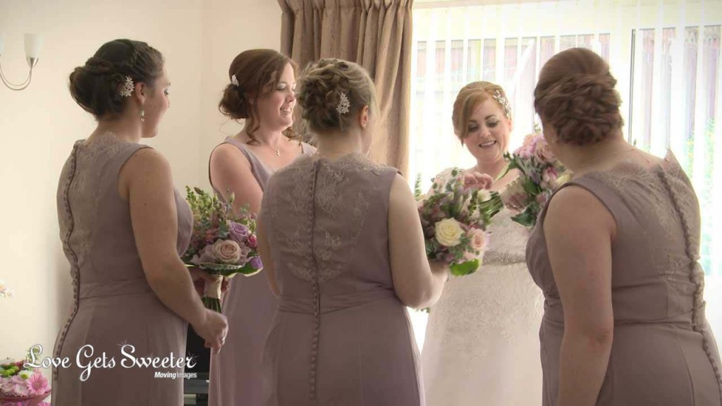 A video still of the bride surrounded by her 4 bridesmaids wearing a dusty pink dress with lace detailing. She passing out the flower bouquets of cream, pinks and dusty purples before they head to the church for her wedding