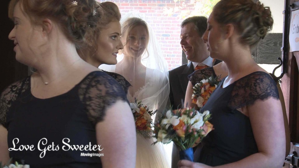 the wedding videographer films through the awaiting bridesmaids wearing navy and holding cream and orange bouquets to see the bride standing with her proud and smiling dad in the doorway to St Johns Church in High Legh