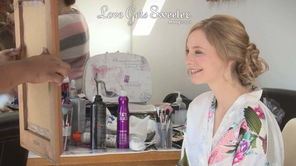 Emma the bride sits in her parents kitchen with a mirror being held in front of her to check her bridal makeup in front of the wedding videographer