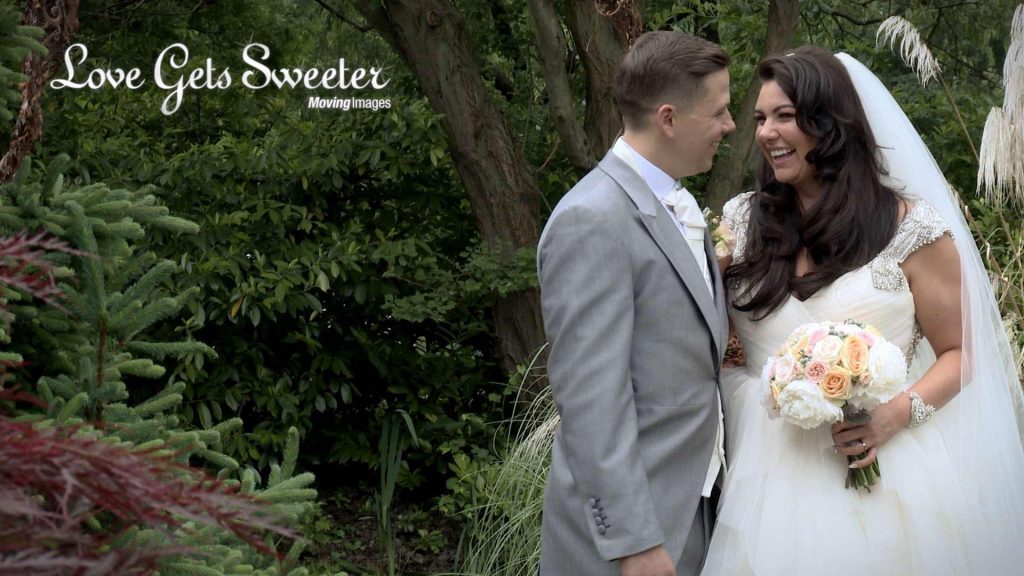 Sherie the bride laughs at her groom as they stand for their photographer and videographer during their wedding day at The Cheshire Hall. She holds a pretty compact bridal bouquet of peach, pink and cream.