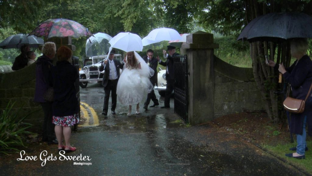 the driver from broughton wedding cars helps the father of the bride and the bride get to the church dry with white umbrellas to stop the rain