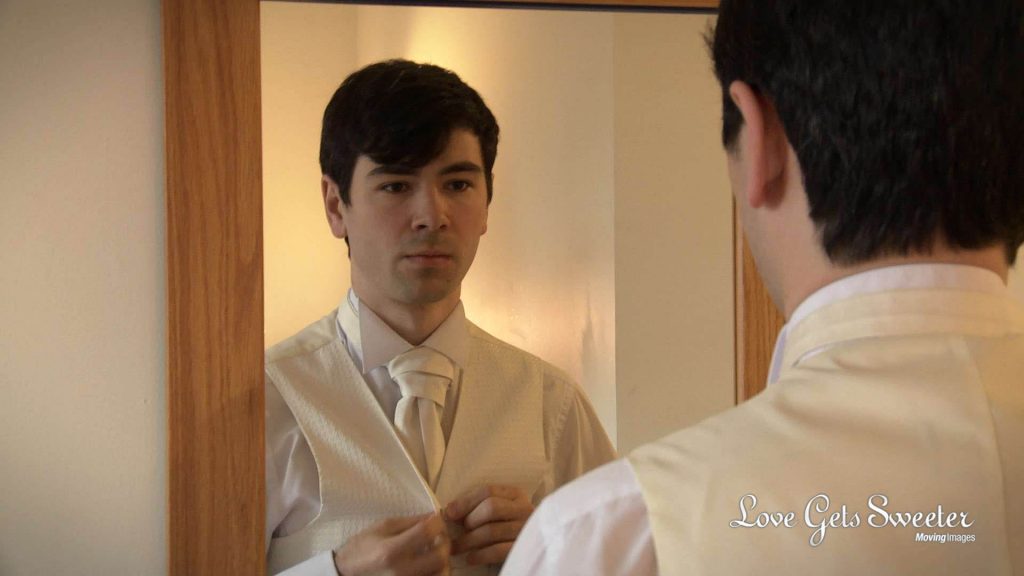 The groom stands in front of a mirror to check his cream wedding tie and waistcoat