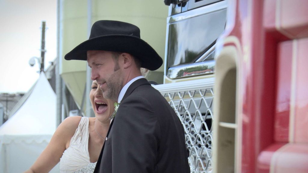 bride and groom laughing on the wedding video while posing in front of the grooms own truck on his family farm
