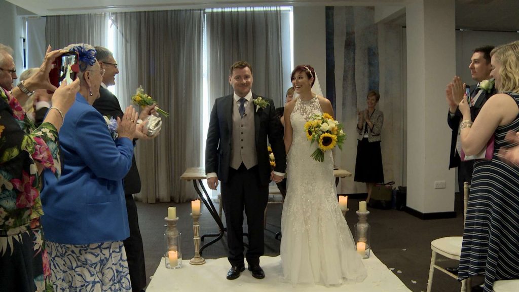 The bride and groom are getting ready to walk down the aisle towards the wedding videographer after being married and look very happy and excited as their wedding guests stand up to cheer. They have a white carpet aisle and she holds an oversized sunflower bouquet