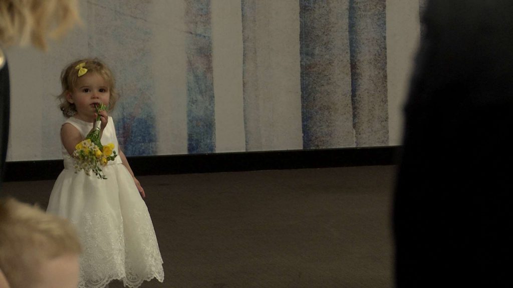 a cute little flower girl wearing white waits and poses at the top of the wedding aisle on the wedding video at her dads wedding at On The 7th
