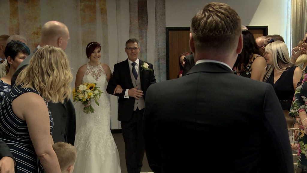 Our short haired bride walks down the aisle with her dad holding a sunflower and white rose bouquet 