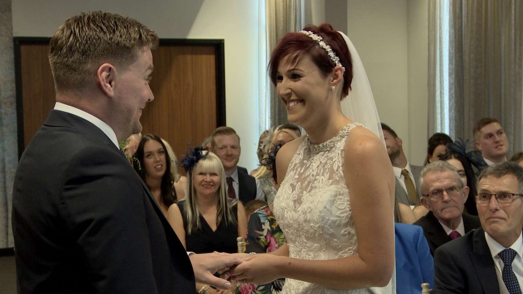 The bride and groom smile and laugh at each other on the wedding video as they exchange their wedding rings during an intimate ceremony at On The 7th in Media City