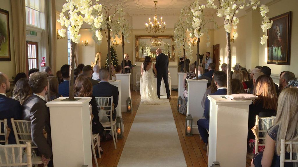 a shot from the back of the ceremony room with a white aisle runner and tall white blossom trees for a winter wedding at Nunsmere Hall in Cheshire