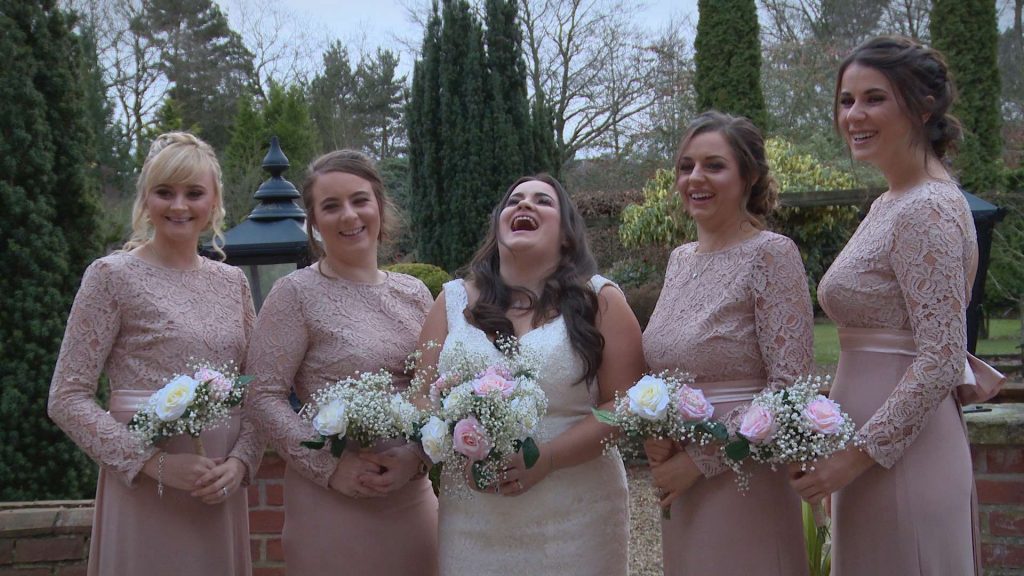 The bride and bridesmaids laugh out loud with their wedding videographer as they have formal photos taken outside nunsmere hall wearing blush pink ASOS bridesmaid dresses and holding faux rose bouquets