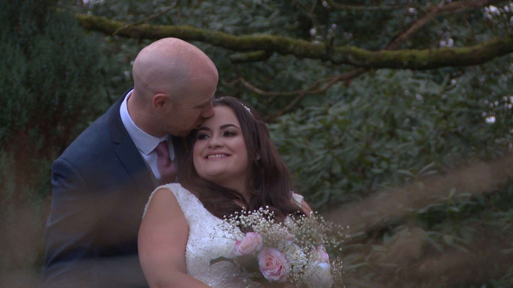 the groom kisses the bride on the forehead as they pose on the bridge at nunsmere hall for their wedding photographer