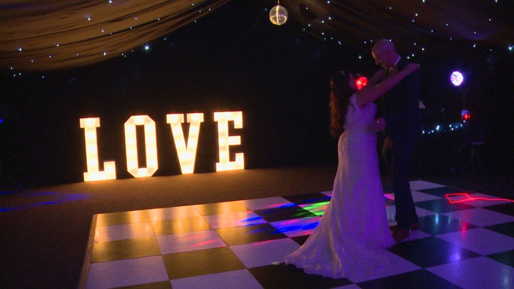 a bride and groom do their first dance for the wedding video on a black and white dance floor with illuminated LOVE letters behind them