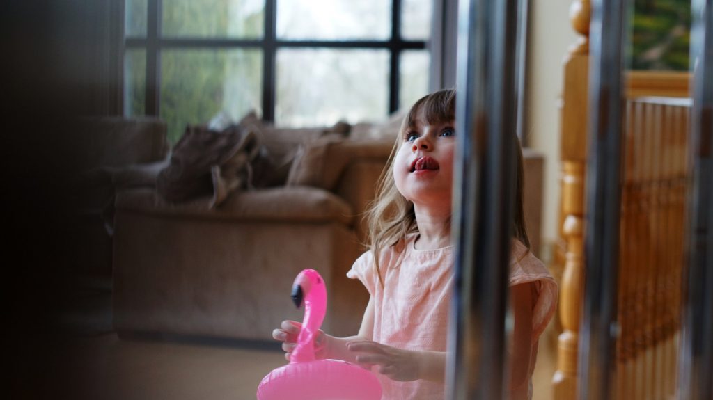 little girl holds a small inflatable pink flamingo in her hand as she looks up past the camera with a little smile