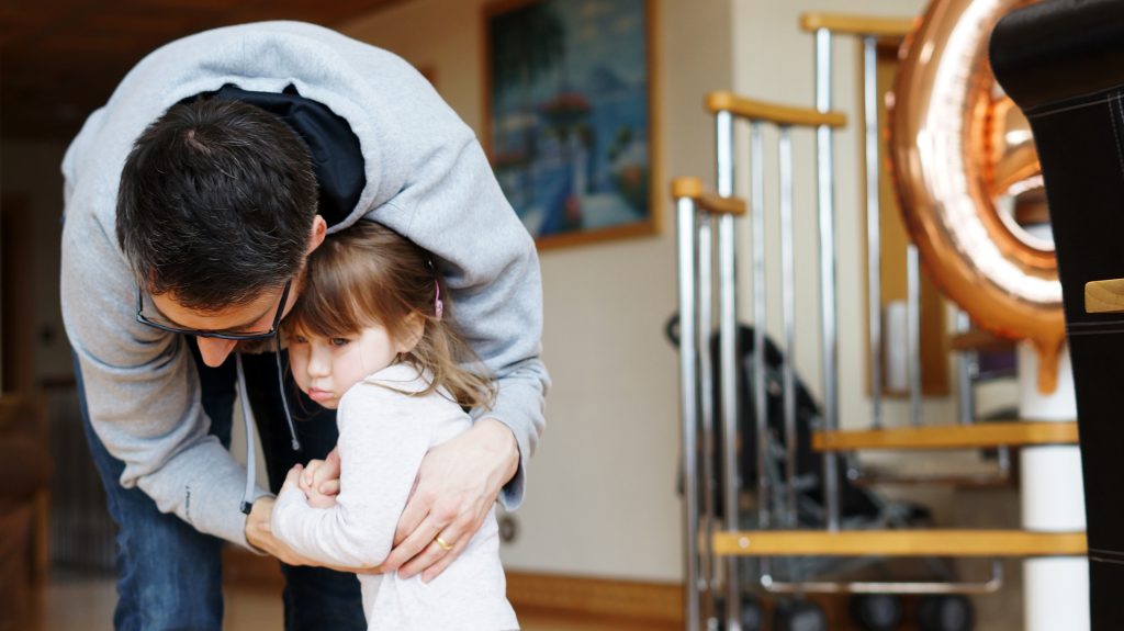 A natural family photo of a young daughter accepts a hug off her Daddy with a pout as he bends down to say goodbye at their holiday home in Penrith
