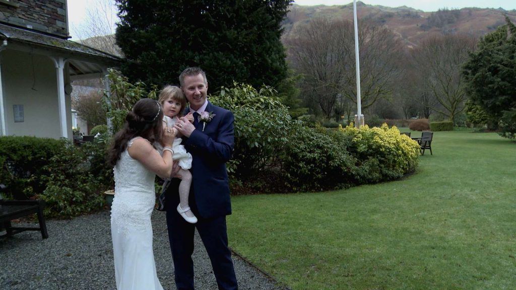the bride and groom try and get their daughter the flower girl to smile for the wedding photographer as they pose in the gardens of the wordsworth hotel with the scenic lake district hills in the background
