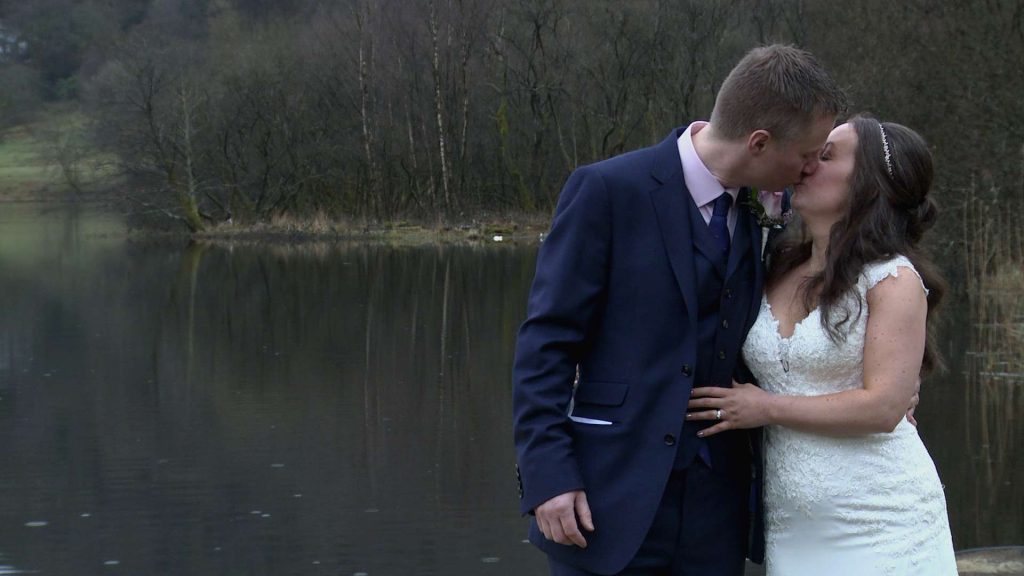 a bride and groom stand in the rain in front of Grasmere lake having a kiss for the wedding photographer
