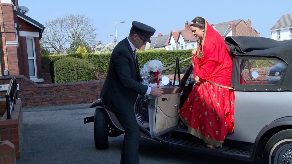 a bride steps out of her traditional silver wedding car outside church for the video wearing a bright red and gold bangladeshi wedding saree dress
