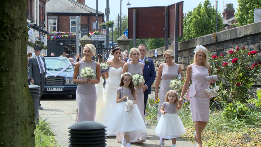 A video still of the bride and her bridal party wearing white and lilac (pale purple) walking down the stone path to make their way in to st johns church in Burscough for the wedding ceremony the bridesmaids are wearing full length lilac purple dresses and holding cream flowers for this summer wedding