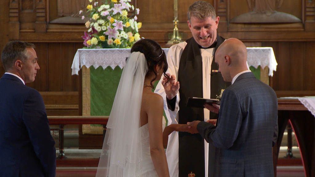 David the priest at St Johns church makes a ring sign with his fingers to show the groom how to push the wedding ring on captured by their wedding videographer Love Gets Sweeter