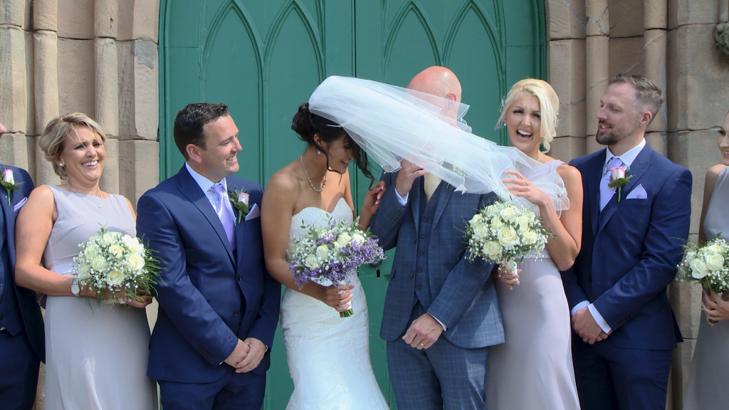 a video still as the brides veil gets blown in front of the grooms face during their wedding photos outside st johns church in burscough