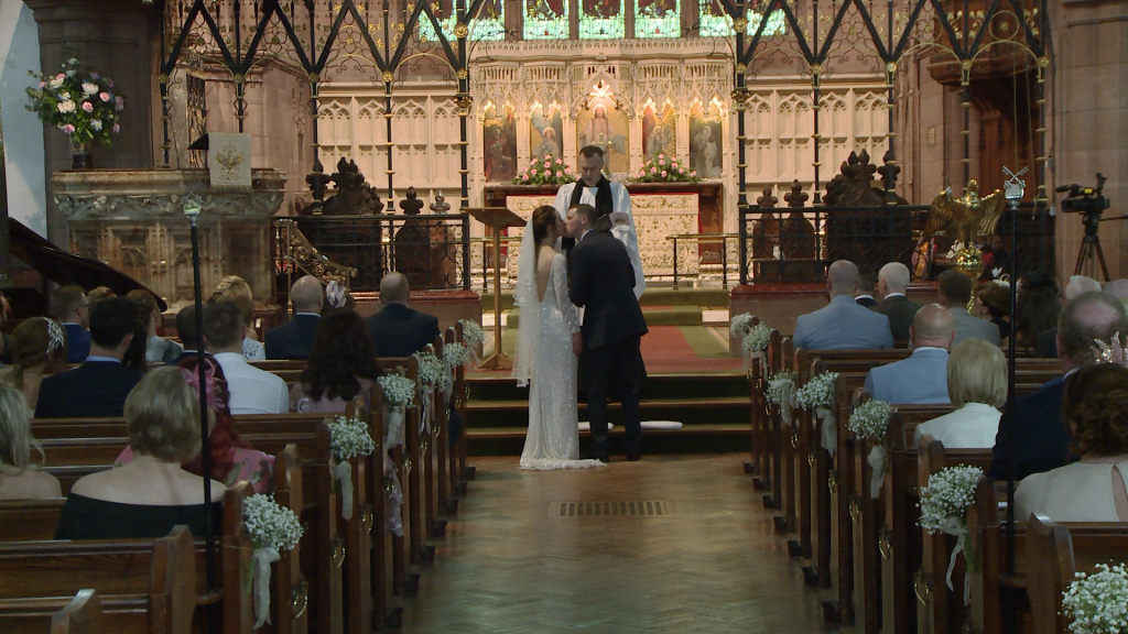 The bride and groom kiss as the priest announces you may kiss the bride in front of family, friends and the wedding videographer at St Peter's Church in Woolton Liverpool