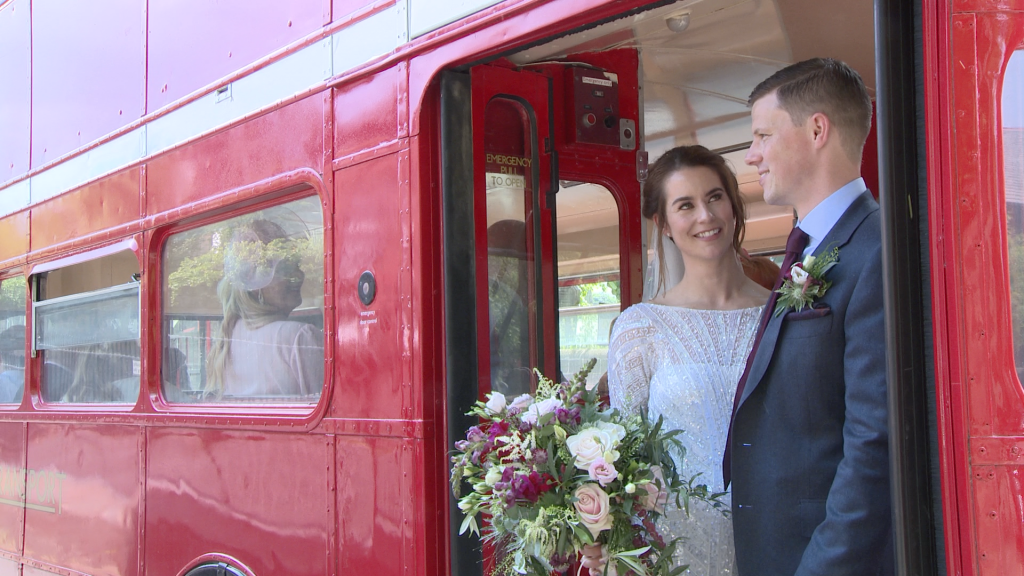 the bride wearing a beaded Eliza Jane Howell dress and holding a rustic purple and pink bouquet and groom wearing a blue grey informal suit and purple tie chat standing on the back of the red london bus after their wedding ceremony at St Peters Church in Woolton Liverpool