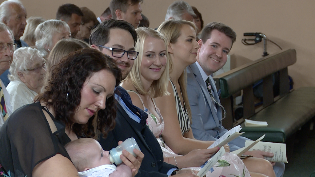 mum dad and godparents smile over at their baby boy having a bottle before his christening at st pauls in Burtonwood