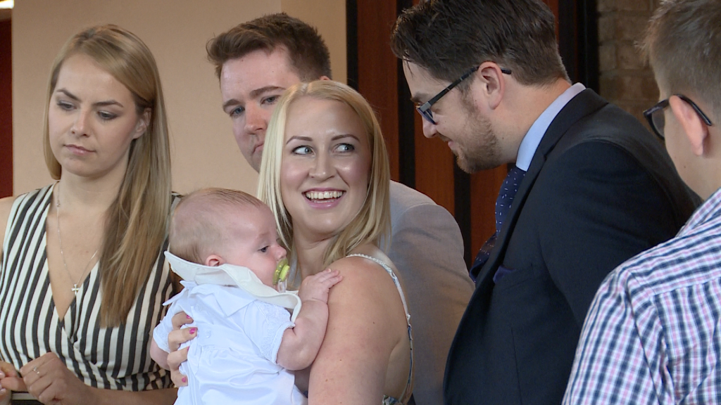 A family photograph of mum smiling at her husband during a christening as she holds her baby in Burtonwood
