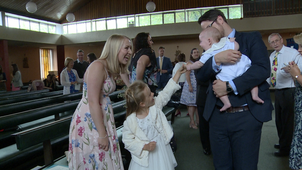 a little girl reaches up to hold a baby boys hand as Mum smiles on at st pauls of the cross in Burtonwood