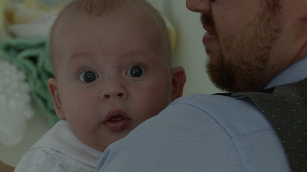 a wide eyed baby boy stares at the videographer over his dads shoulders