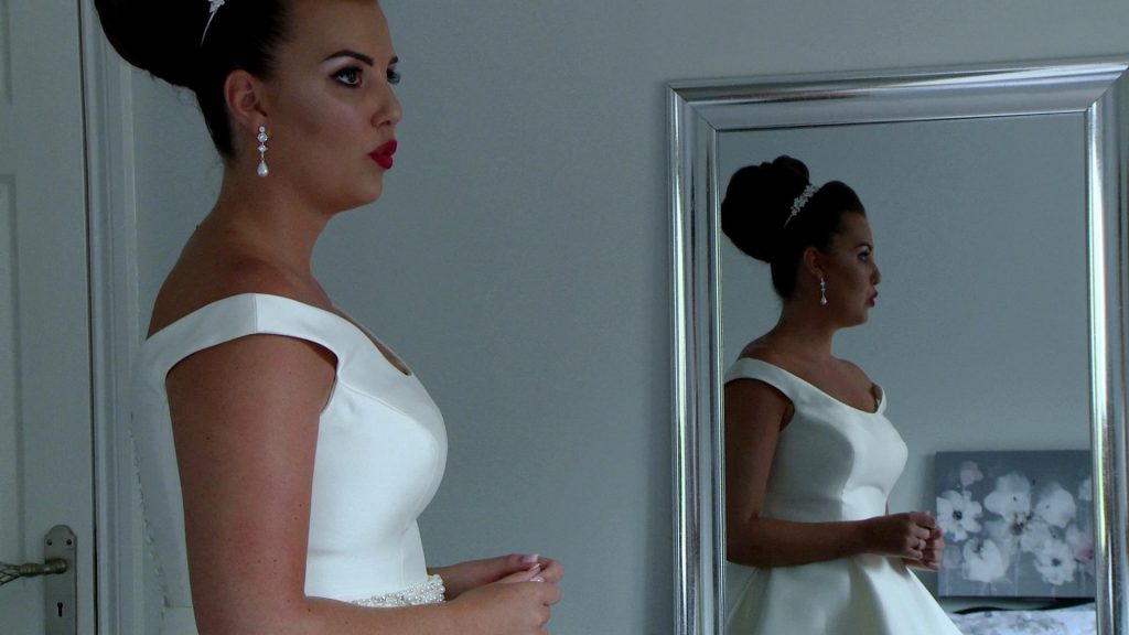 a classic old hollywood style bride takes a deep breath pouting her red lips with her reflection in the mirror behind her on the wedding video