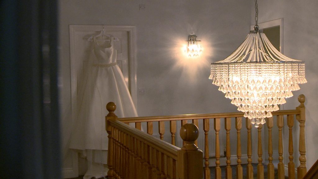a stunning white satin Ellis bridal dress hangs on a door frame by an elegant chandelier by the staircase
